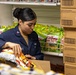 A U.S. military service member in navy blue utility uniform is stocking or organizing snack food items in what appears to be a military exchange or commissary store. She is handling chip bags near stacked cardboard boxes of Doritos, with Calbee Crispy Potato chip bags visible on a wire shelf behind her. The setting suggests a retail or supply storage area on a military installation.