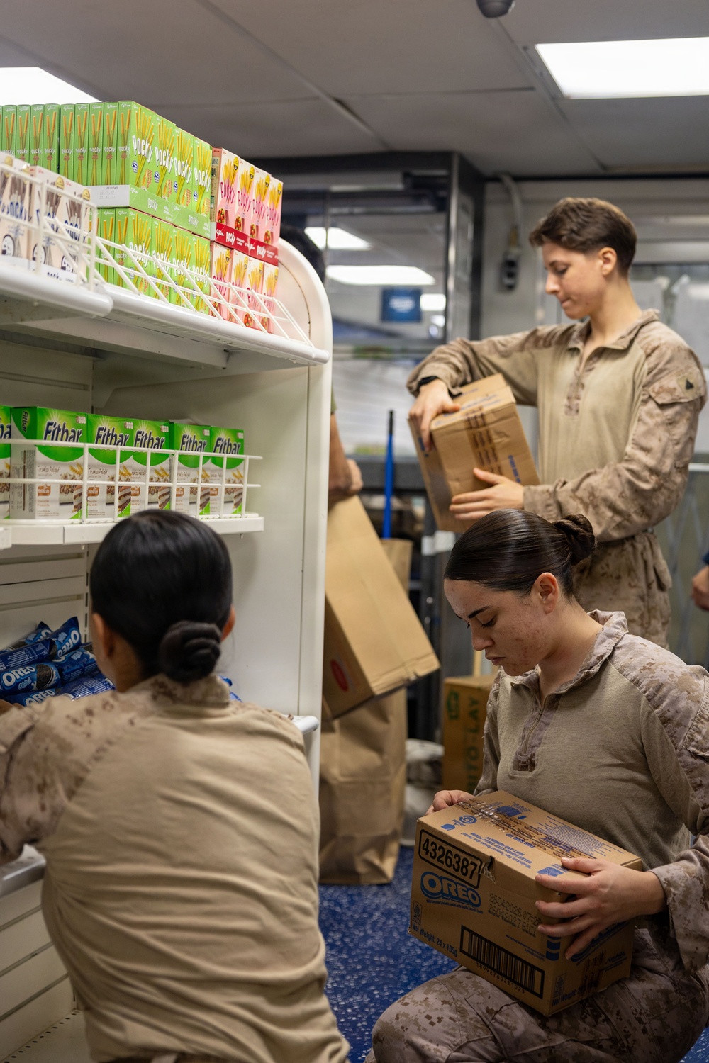 Three individuals in military camouflage uniforms are restocking shelves in what appears to be a small military exchange or convenience store. They are handling cardboard boxes and placing snack products onto white retail shelving units. Visible products include Oreo cookies, Pocky snack sticks, and Fitbar snack bars.