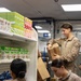 Three individuals in military camouflage uniforms are restocking shelves in what appears to be a small military exchange or convenience store. They are handling cardboard boxes and placing snack products onto white retail shelving units. Visible products include Oreo cookies, Pocky snack sticks, and Fitbar snack bars.