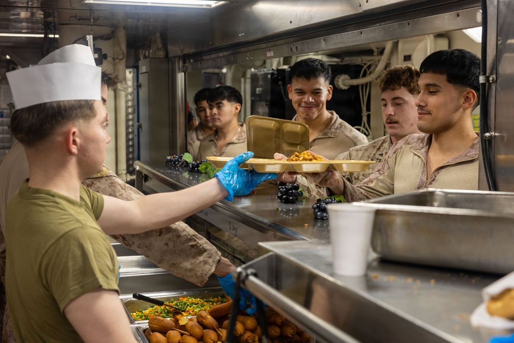 A military mess hall scene showing a food service worker in a white cook's hat and blue gloves serving meals to a line of personnel in camouflage uniforms. The setting appears to be aboard a naval vessel or military installation, with stainless steel serving equipment and institutional lighting visible. Personnel are receiving food trays containing what appears to be a hot meal with side items.