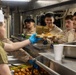 A military mess hall scene showing a food service worker in a white cook's hat and blue gloves serving meals to a line of personnel in camouflage uniforms. The setting appears to be aboard a naval vessel or military installation, with stainless steel serving equipment and institutional lighting visible. Personnel are receiving food trays containing what appears to be a hot meal with side items.