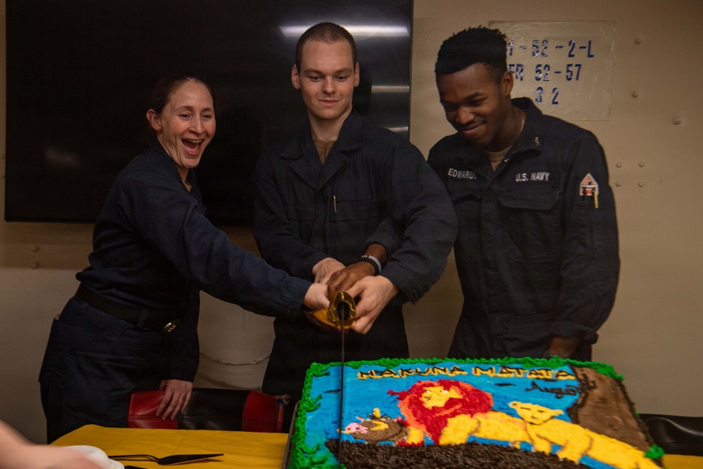 Three U.S. Navy personnel in dark blue coveralls are cutting a decorated cake aboard what appears to be a naval vessel. The cake features a Lion King theme with 'Hakuna Matata' written in frosting and depicts characters from the film. The sailors appear to be in good spirits, with one woman laughing openly while two male sailors assist in cutting the cake with what appears to be a ceremonial sword or knife.