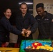 Three U.S. Navy personnel in dark blue coveralls are cutting a decorated cake aboard what appears to be a naval vessel. The cake features a Lion King theme with 'Hakuna Matata' written in frosting and depicts characters from the film. The sailors appear to be in good spirits, with one woman laughing openly while two male sailors assist in cutting the cake with what appears to be a ceremonial sword or knife.