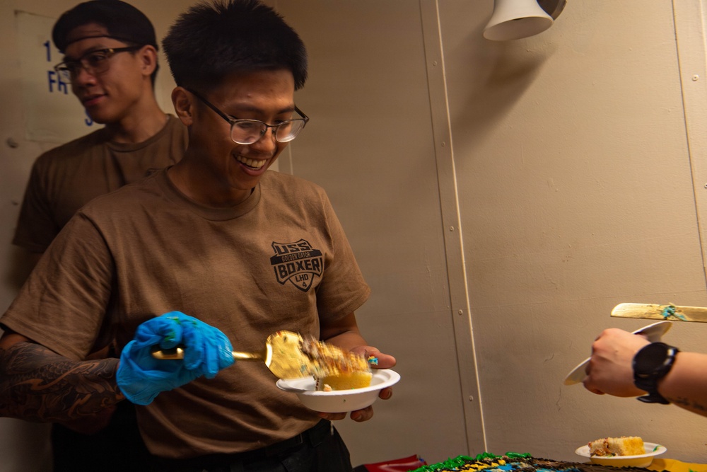 A smiling young man wearing a USS Boxer LHD-4 t-shirt and blue latex gloves is serving cake from a decorated sheet cake. He is holding a plate with a slice of cake and a serving spatula. A second person is visible in the background, and another individual's hand extends from the right side of the frame holding a spatula.