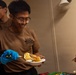 A smiling young man wearing a USS Boxer LHD-4 t-shirt and blue latex gloves is serving cake from a decorated sheet cake. He is holding a plate with a slice of cake and a serving spatula. A second person is visible in the background, and another individual's hand extends from the right side of the frame holding a spatula.