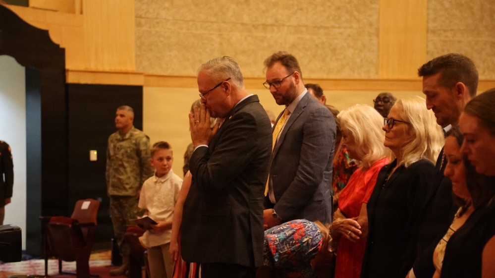 A formal indoor ceremony or service is taking place in what appears to be a large hall or auditorium. A group of civilians and at least one military personnel are standing, with the central figure appearing to be in a moment of prayer or emotional reflection with hands raised to his face. The setting and attire suggest an official or solemn occasion such as a memorial, retirement ceremony, or formal military-affiliated event.