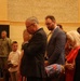 A formal indoor ceremony or service is taking place in what appears to be a large hall or auditorium. A group of civilians and at least one military personnel are standing, with the central figure appearing to be in a moment of prayer or emotional reflection with hands raised to his face. The setting and attire suggest an official or solemn occasion such as a memorial, retirement ceremony, or formal military-affiliated event.