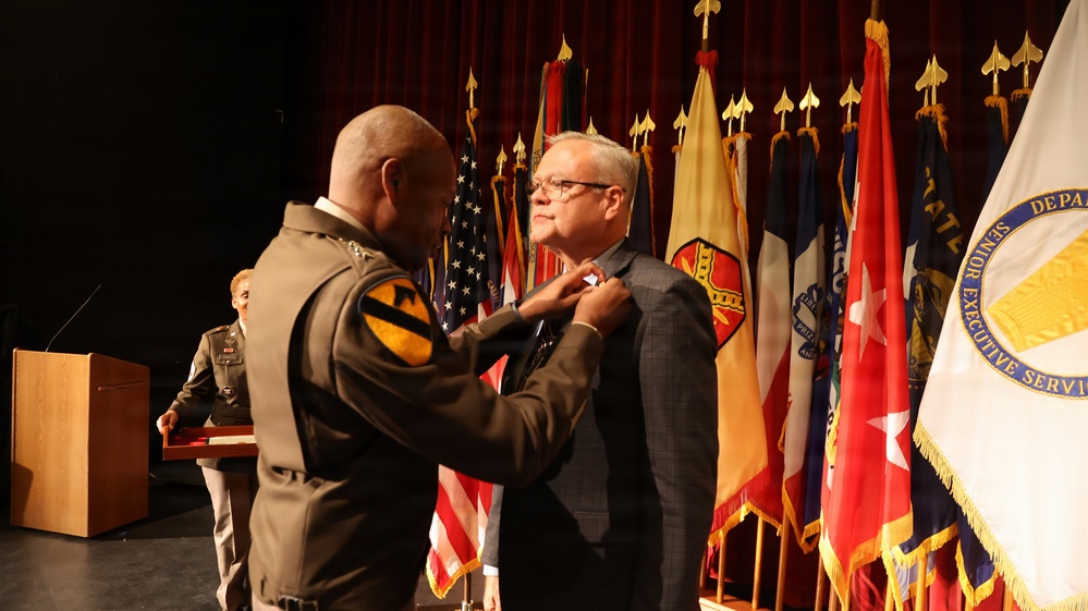 A military officer in Army dress uniform is pinning a medal or award onto a civilian man in a suit jacket during a formal ceremony. The event takes place on a stage with multiple military and organizational flags displayed in the background. A podium and another uniformed soldier are visible in the background.