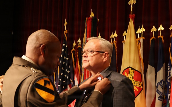 A military officer in Army dress uniform is pinning a medal or award onto a civilian man in a suit jacket during a formal ceremony. The event takes place on a stage with multiple military and organizational flags displayed in the background. A podium and another uniformed soldier are visible in the background.