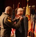 A military officer in Army dress uniform is pinning a medal or award onto a civilian man in a suit jacket during a formal ceremony. The event takes place on a stage with multiple military and organizational flags displayed in the background. A podium and another uniformed soldier are visible in the background.