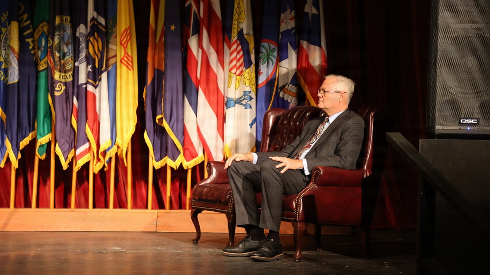 An older man in a gray suit and tie is seated in a tufted leather armchair on a stage, appearing to be participating in a formal event or interview. Behind him is a display of numerous flags representing U.S. states and territories, arranged on a draped backdrop. A QSC speaker is visible on the right side of the stage.