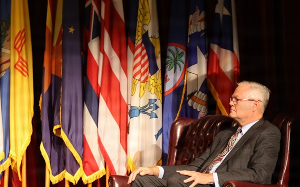 An older man in a gray suit and tie is seated in a tufted leather armchair on a stage, appearing to be participating in a formal event or interview. Behind him is a display of numerous flags representing U.S. states and territories, arranged on a draped backdrop. A QSC speaker is visible on the right side of the stage.