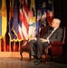An older man in a gray suit and tie is seated in a tufted leather armchair on a stage, appearing to be participating in a formal event or interview. Behind him is a display of numerous flags representing U.S. states and territories, arranged on a draped backdrop. A QSC speaker is visible on the right side of the stage.