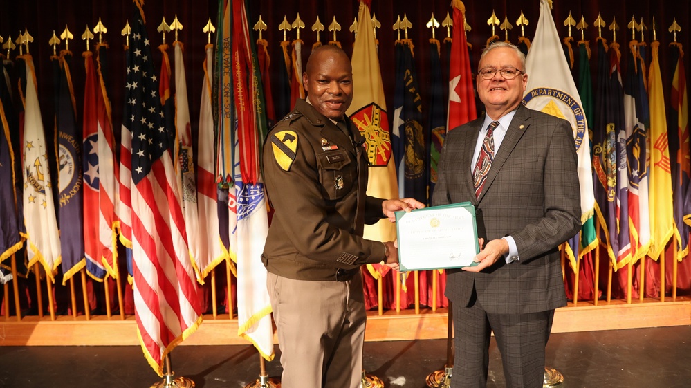 Two individuals are posing together on a stage while jointly holding a certificate or award document. One person is wearing a U.S. Army uniform with a 1st Cavalry Division patch, while the other is dressed in civilian business attire. The ceremony appears to be taking place in a formal military setting surrounded by numerous military and state flags.
