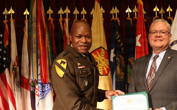 Two individuals are posing together on a stage while jointly holding a certificate or award document. One person is wearing a U.S. Army uniform with a 1st Cavalry Division patch, while the other is dressed in civilian business attire. The ceremony appears to be taking place in a formal military setting surrounded by numerous military and state flags.