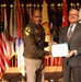 Two individuals are posing together on a stage while jointly holding a certificate or award document. One person is wearing a U.S. Army uniform with a 1st Cavalry Division patch, while the other is dressed in civilian business attire. The ceremony appears to be taking place in a formal military setting surrounded by numerous military and state flags.