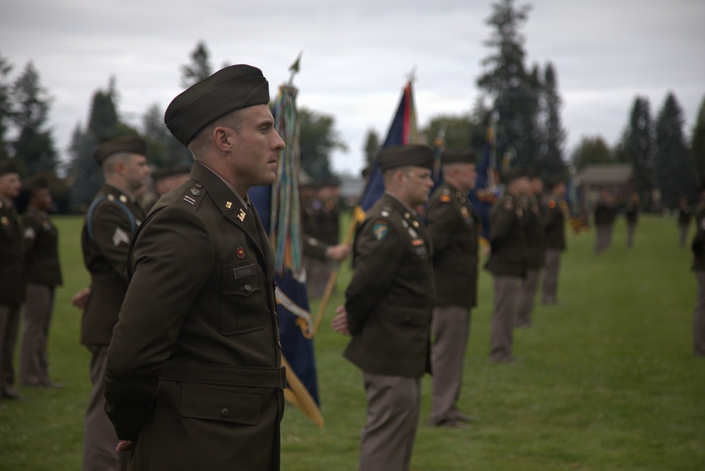 A formal U.S. Army military ceremony is taking place on a grass field, with soldiers in dress green uniforms standing in formation. Multiple color guard members are visible holding unit guidons and flags in the background. The foreground features a soldier wearing a distinctive black beret and officer insignia standing at attention.