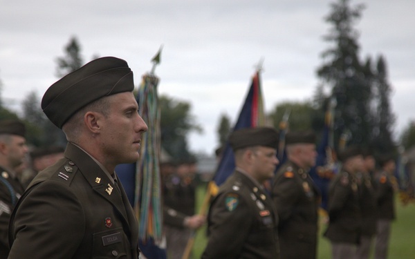 A formal U.S. Army military ceremony is taking place on a grass field, with soldiers in dress green uniforms standing in formation. Multiple color guard members are visible holding unit guidons and flags in the background. The foreground features a soldier wearing a distinctive black beret and officer insignia standing at attention.