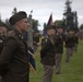 A formal U.S. Army military ceremony is taking place on a grass field, with soldiers in dress green uniforms standing in formation. Multiple color guard members are visible holding unit guidons and flags in the background. The foreground features a soldier wearing a distinctive black beret and officer insignia standing at attention.