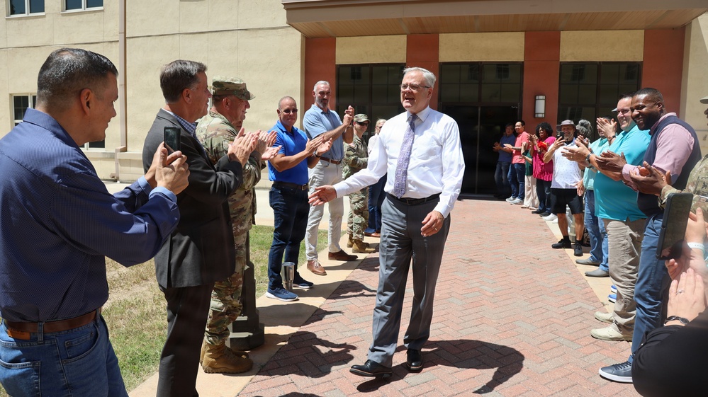 A man in a white dress shirt and gray slacks walks through a corridor of applauding people outside a tan/brick institutional building. The scene appears to be a farewell or recognition ceremony, with a mix of civilians and at least one military personnel in camouflage uniform forming two lines to honor the central figure. The atmosphere is celebratory, with people smiling, clapping, and some recording the moment on smartphones.