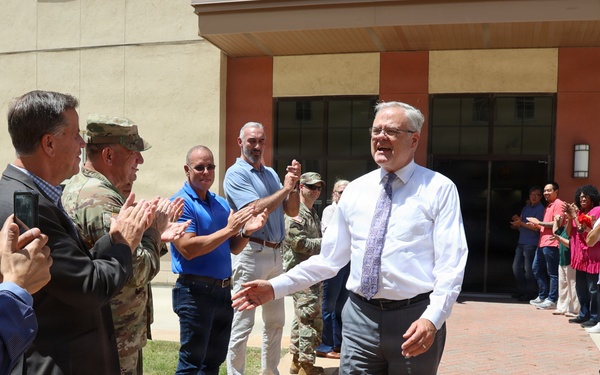 A man in a white dress shirt and gray slacks walks through a corridor of applauding people outside a tan/brick institutional building. The scene appears to be a farewell or recognition ceremony, with a mix of civilians and at least one military personnel in camouflage uniform forming two lines to honor the central figure. The atmosphere is celebratory, with people smiling, clapping, and some recording the moment on smartphones.