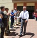 A man in a white dress shirt and gray slacks walks through a corridor of applauding people outside a tan/brick institutional building. The scene appears to be a farewell or recognition ceremony, with a mix of civilians and at least one military personnel in camouflage uniform forming two lines to honor the central figure. The atmosphere is celebratory, with people smiling, clapping, and some recording the moment on smartphones.