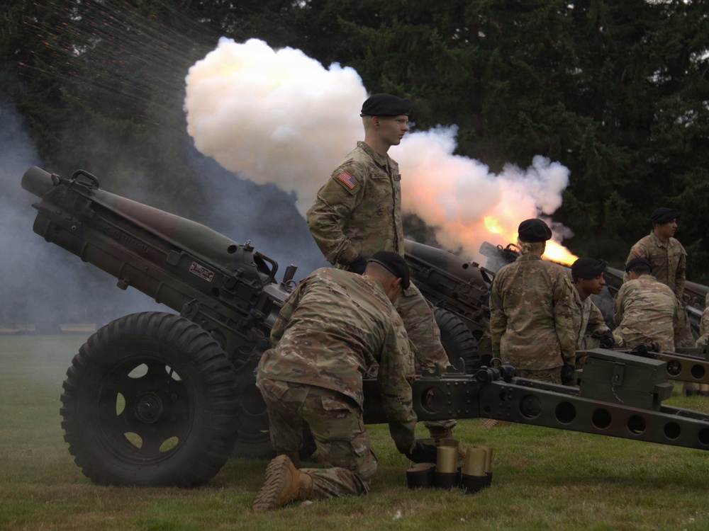 U.S. Army soldiers in camouflage uniforms and black berets are operating artillery pieces during what appears to be a ceremonial cannon salute or training exercise. At least two howitzers are visible, with one actively firing, producing a large cloud of white smoke and visible muzzle flash. The setting is an outdoor grassy area with trees in the background.