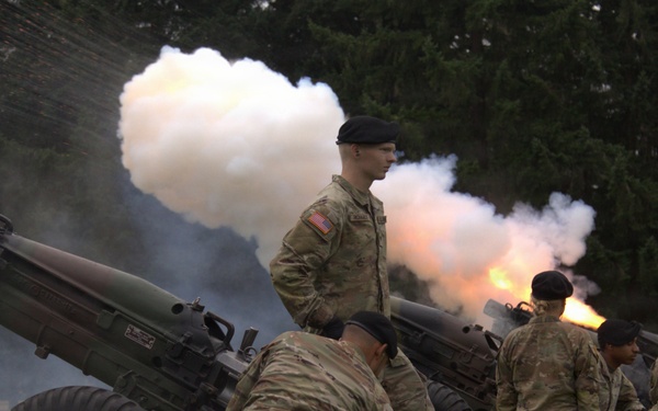 U.S. Army soldiers in camouflage uniforms and black berets are operating artillery pieces during what appears to be a ceremonial cannon salute or training exercise. At least two howitzers are visible, with one actively firing, producing a large cloud of white smoke and visible muzzle flash. The setting is an outdoor grassy area with trees in the background.