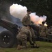 U.S. Army soldiers in camouflage uniforms and black berets are operating artillery pieces during what appears to be a ceremonial cannon salute or training exercise. At least two howitzers are visible, with one actively firing, producing a large cloud of white smoke and visible muzzle flash. The setting is an outdoor grassy area with trees in the background.