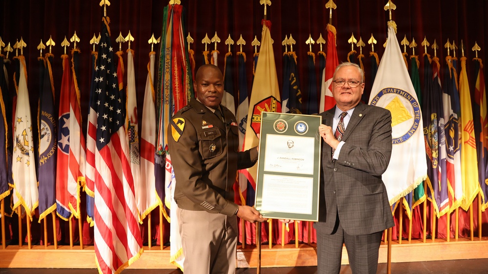Two individuals are posing together on a stage holding a framed certificate or award. One person is wearing a U.S. Army military uniform with a 1st Cavalry Division patch, while the other is in civilian business attire. The ceremony appears to be an official military recognition event, with the certificate bearing the name 'J. Randall Robinson'.