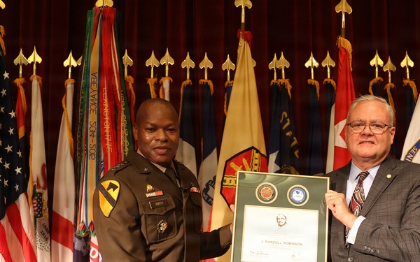 Two individuals are posing together on a stage holding a framed certificate or award. One person is wearing a U.S. Army military uniform with a 1st Cavalry Division patch, while the other is in civilian business attire. The ceremony appears to be an official military recognition event, with the certificate bearing the name 'J. Randall Robinson'.