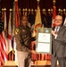 Two individuals are posing together on a stage holding a framed certificate or award. One person is wearing a U.S. Army military uniform with a 1st Cavalry Division patch, while the other is in civilian business attire. The ceremony appears to be an official military recognition event, with the certificate bearing the name 'J. Randall Robinson'.