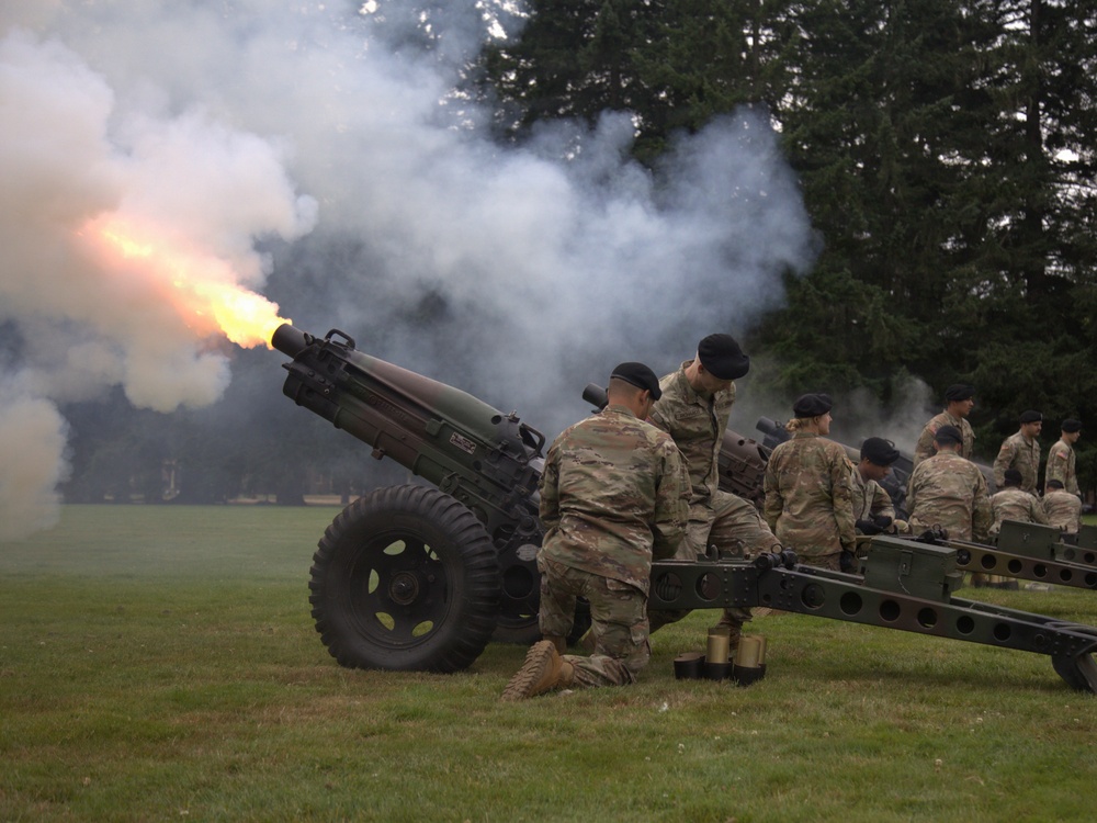 Military personnel in camouflage uniforms and black berets are conducting a ceremonial cannon salute on a grass field. At least one cannon is actively firing, producing a visible muzzle flash and large plume of white smoke. Multiple artillery pieces and soldiers are visible in a line, suggesting a coordinated multi-gun salute.