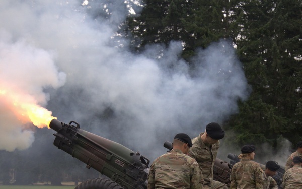 Military personnel in camouflage uniforms and black berets are conducting a ceremonial cannon salute on a grass field. At least one cannon is actively firing, producing a visible muzzle flash and large plume of white smoke. Multiple artillery pieces and soldiers are visible in a line, suggesting a coordinated multi-gun salute.