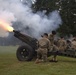 Military personnel in camouflage uniforms and black berets are conducting a ceremonial cannon salute on a grass field. At least one cannon is actively firing, producing a visible muzzle flash and large plume of white smoke. Multiple artillery pieces and soldiers are visible in a line, suggesting a coordinated multi-gun salute.