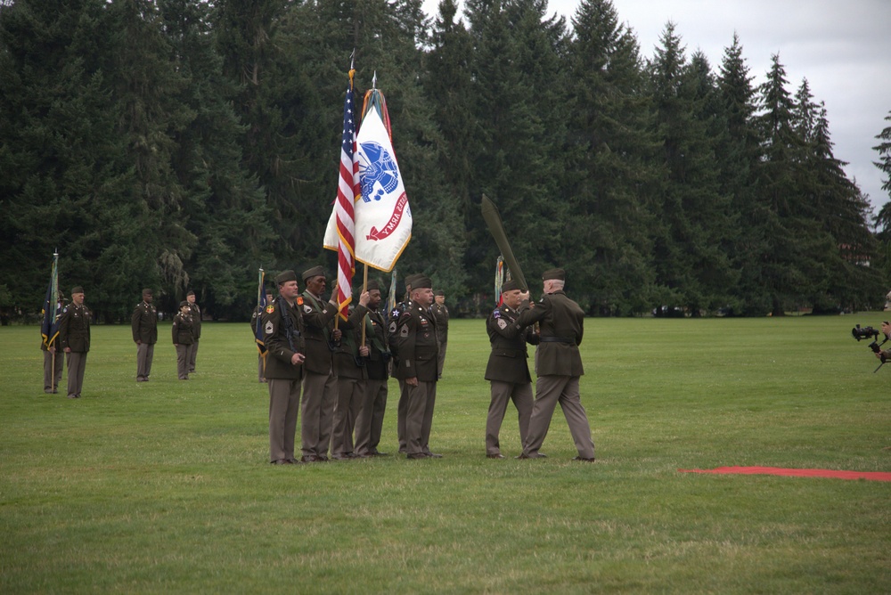 A formal U.S. Army military ceremony is taking place on a grass parade field, likely a change of command or activation/inactivation ceremony. Soldiers in Army Service Uniforms (green Class A uniforms) are participating in a colors/guidon transfer, with the U.S. flag and a unit flag prominently displayed. The setting appears to be a military installation surrounded by tall evergreen trees, consistent with the Pacific Northwest region.