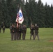 A formal U.S. Army military ceremony is taking place on a grass parade field, likely a change of command or activation/inactivation ceremony. Soldiers in Army Service Uniforms (green Class A uniforms) are participating in a colors/guidon transfer, with the U.S. flag and a unit flag prominently displayed. The setting appears to be a military installation surrounded by tall evergreen trees, consistent with the Pacific Northwest region.