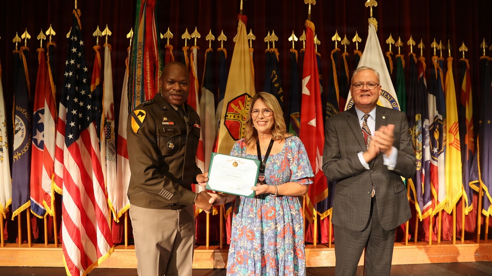 A formal military award ceremony taking place on a stage with numerous state and military flags displayed in the background. A U.S. Army officer in uniform is presenting a framed certificate to a woman in a floral dress, while a civilian man in a suit applauds to the right. The setting appears to be an official government or military installation auditorium.
