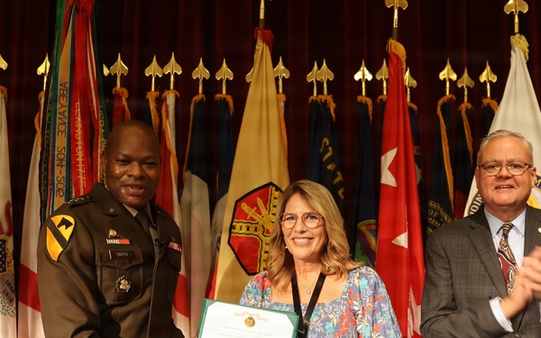 A formal military award ceremony taking place on a stage with numerous state and military flags displayed in the background. A U.S. Army officer in uniform is presenting a framed certificate to a woman in a floral dress, while a civilian man in a suit applauds to the right. The setting appears to be an official government or military installation auditorium.