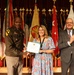 A formal military award ceremony taking place on a stage with numerous state and military flags displayed in the background. A U.S. Army officer in uniform is presenting a framed certificate to a woman in a floral dress, while a civilian man in a suit applauds to the right. The setting appears to be an official government or military installation auditorium.