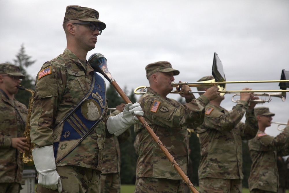 A U.S. Army military band is performing at an outdoor ceremony, likely on a military installation. Soldiers in MultiCam/OCP camouflage uniforms are playing brass instruments including trombones and what appears to be a saxophone. A drum major or color bearer in the foreground holds a ceremonial mace and a blue unit guidon banner.