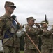 A U.S. Army military band is performing at an outdoor ceremony, likely on a military installation. Soldiers in MultiCam/OCP camouflage uniforms are playing brass instruments including trombones and what appears to be a saxophone. A drum major or color bearer in the foreground holds a ceremonial mace and a blue unit guidon banner.
