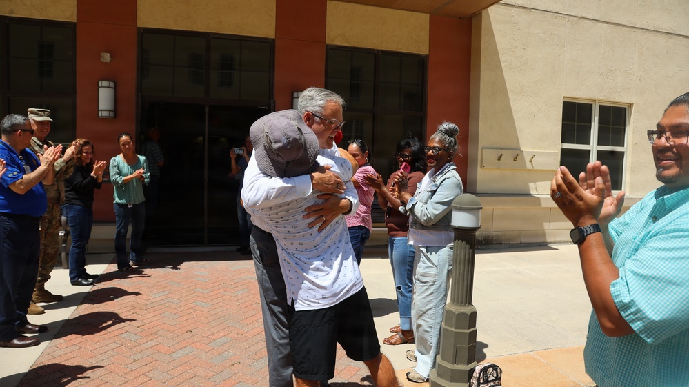 Two individuals are sharing an emotional embrace in front of a building entrance while a group of onlookers applauds around them. The scene appears to be a celebratory or reunion moment, possibly at a government or military facility given the presence of a person in military uniform. The crowd's applause and smiling expressions suggest a positive, significant occasion.