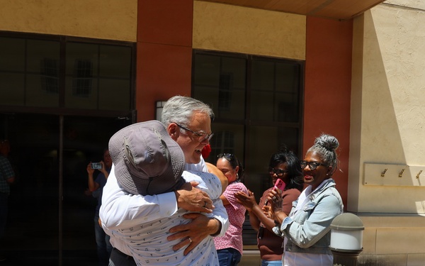 Two individuals are sharing an emotional embrace in front of a building entrance while a group of onlookers applauds around them. The scene appears to be a celebratory or reunion moment, possibly at a government or military facility given the presence of a person in military uniform. The crowd's applause and smiling expressions suggest a positive, significant occasion.