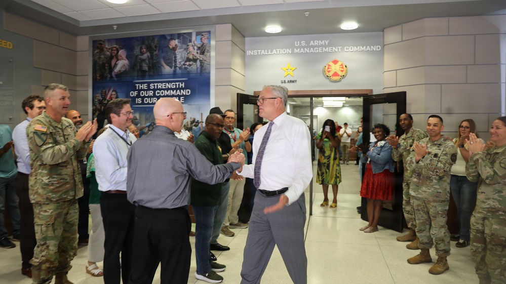 A formal reception or farewell ceremony is taking place in the lobby of the U.S. Army Installation Management Command headquarters. Two civilians in the center are shaking hands while a crowd of military personnel in Army Combat Uniforms and civilians line both sides, applauding. The setting suggests an official recognition, welcome, or departure event.