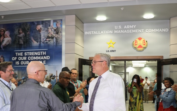 A formal reception or farewell ceremony is taking place in the lobby of the U.S. Army Installation Management Command headquarters. Two civilians in the center are shaking hands while a crowd of military personnel in Army Combat Uniforms and civilians line both sides, applauding. The setting suggests an official recognition, welcome, or departure event.