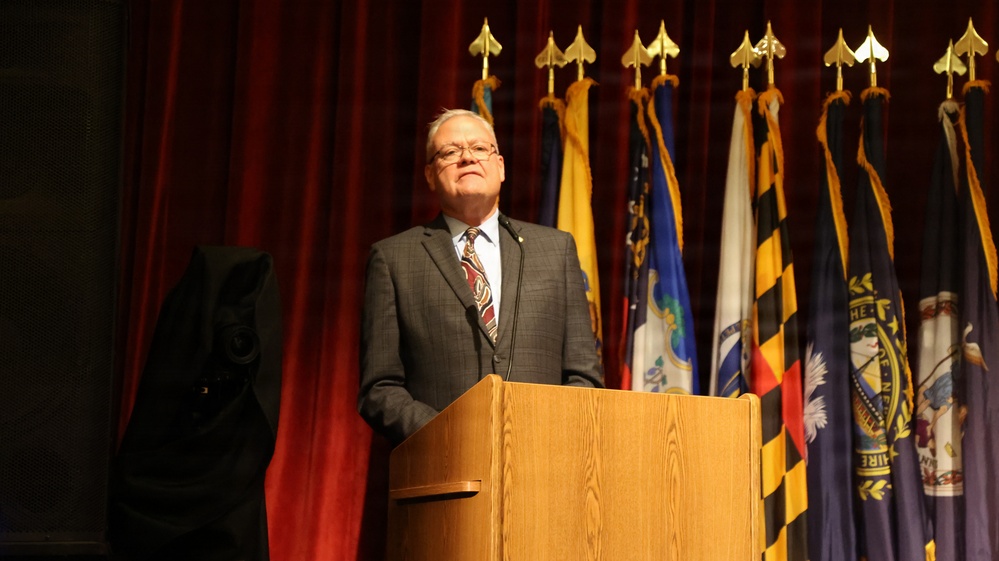 A man in a gray suit and patterned tie stands at a wooden podium, appearing to deliver a speech or address. Behind him, a row of flags on staffs with gold finials is displayed against a red curtain backdrop. The setting appears to be a formal or official ceremony.