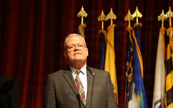 A man in a gray suit and patterned tie stands at a wooden podium, appearing to deliver a speech or address. Behind him, a row of flags on staffs with gold finials is displayed against a red curtain backdrop. The setting appears to be a formal or official ceremony.