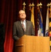A man in a gray suit and patterned tie stands at a wooden podium, appearing to deliver a speech or address. Behind him, a row of flags on staffs with gold finials is displayed against a red curtain backdrop. The setting appears to be a formal or official ceremony.