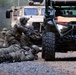 Military personnel in camouflage uniforms are conducting a tactical training exercise on a gravel road in a wooded area. Soldiers are positioned in various combat stances including prone firing position and crouching, with weapons drawn. The scene includes multiple military vehicles including a tan MRAP/JLTV and an olive-drab utility side-by-side vehicle (UTV) with an orange panel visible.
